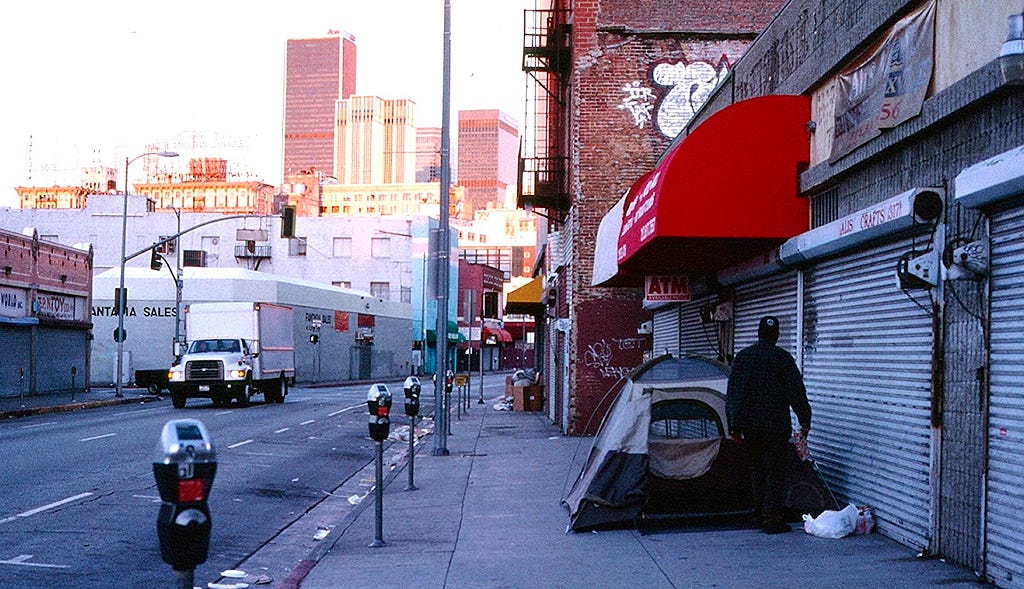 A photograph of Skid Row back in 2010 shows a city street with a truck passing by, a person standing near their tent on the sidewalk, and buildings in downtown Los Angeles in the background.