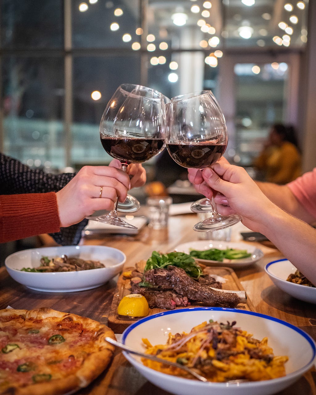 Inside a restaurant two people cheer with red wine. The wine glasses are the focus and only see the hands of the people as they cheer the glasses from either side of the table.