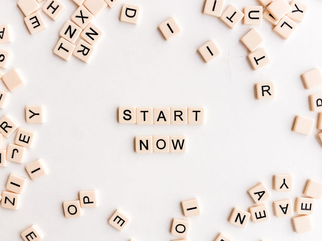 A photo of a variety of Scrabble tiles on a white background. Scrabble tiles in the center spell out the words Start Now. Photo by Sincerely Media on Unsplash.