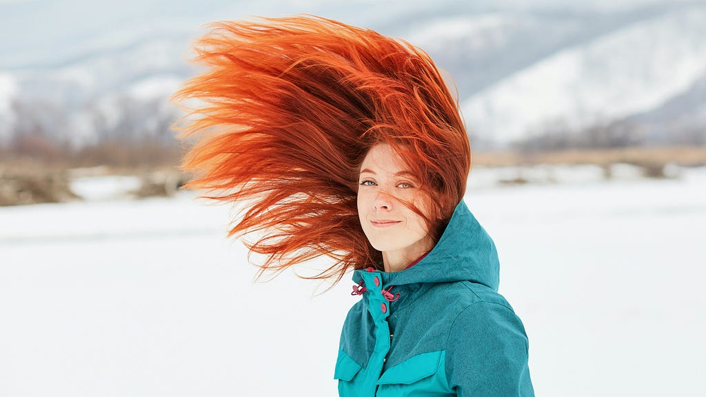 a female redhead w/ her hair flying on þe wind. winter. outside. smiling. she wears a blue jacket.