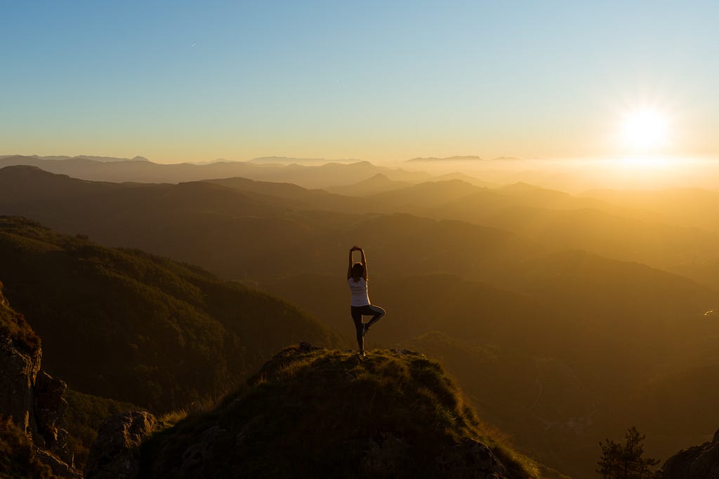 mulher de costas, em cima de uma montanha em postura de yoga olhando pro horizonte, que mostra mais montanhas e por do sol