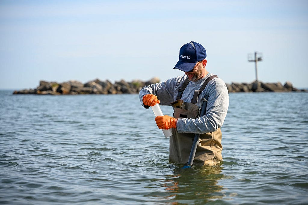 Man in waders stands waist deep in lake water closing the cap of a sample bottle.