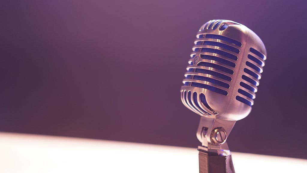 photo of an old-style microphone with silver metal outer and open lines in the structure of the device. The background is dark and blurry, with a clearly lit blurry spotlight shining where the mic is placed.