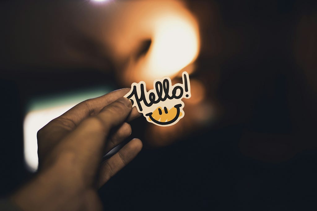 A male hand holds a smiley face sticker with the word “hello” on it.