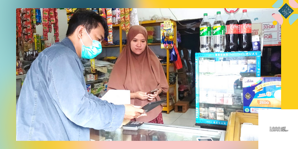 A photo showing a male field officer helping a female agent onboard to the GrabKios in a small snack shop. The officer wears a denim blue jacket, and the agent wears a brown headscarf. She stands behind the counter at her shop and holds a phone, looking at the tablet the officer is holding.