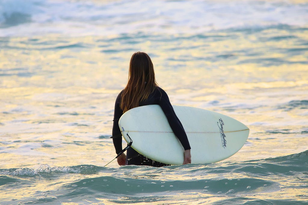 A woman in a wetsuit holding a surf board as she wades into the water.