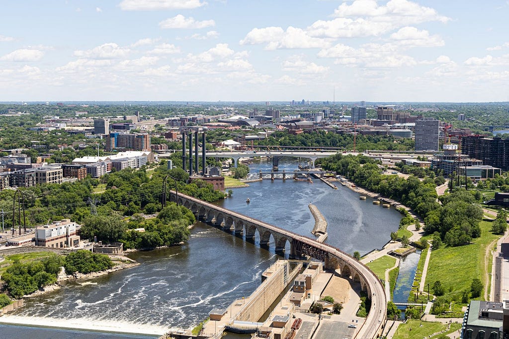 An aerial photograph of Minneapolis-Saint Paul, Minnesota shows the city’s river being crossed by bridges and flanked by buildings and trees.