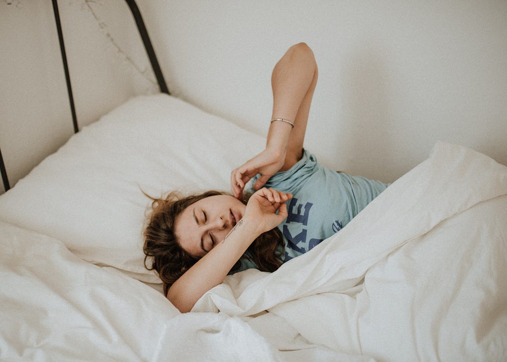 Photo of a woman in a blue t-shirt lying in a bed with white bedding. She has her eyes closed and her arms outstretched.