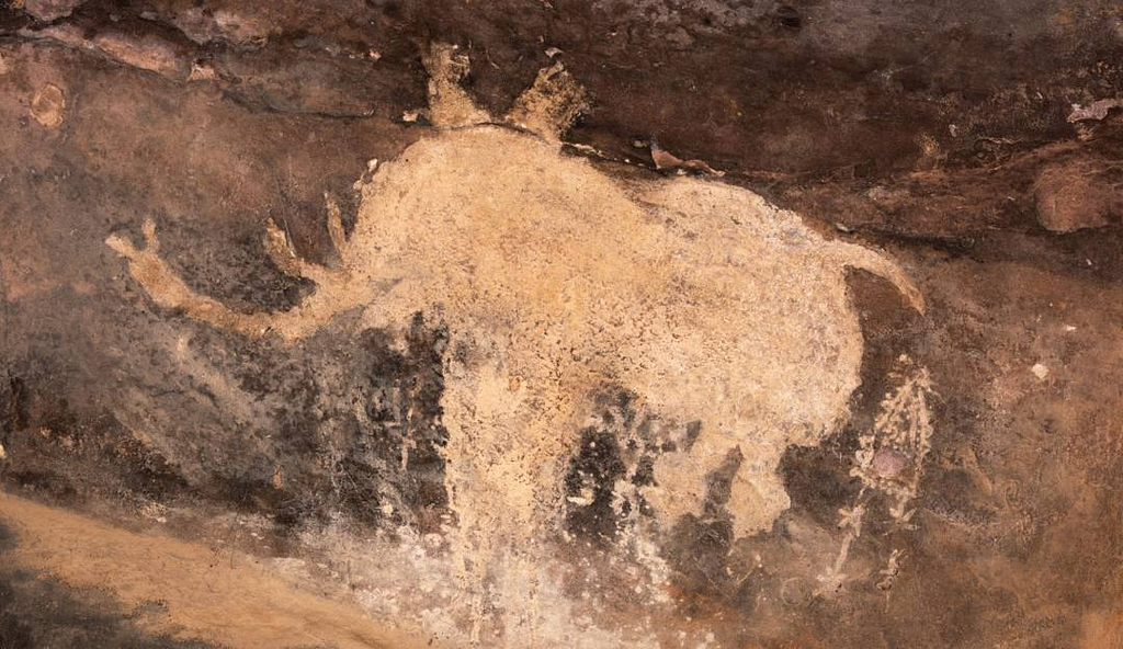 Ancient cave painting featuring a large, faded depiction of an animal, possibly a bison or a wild bull, painted in a light pigment against a dark rock surface. The rough texture of the rock and the weathered appearance of the artwork indicate its prehistoric origins. The figure’s outline is partially eroded, but the shape of the animal’s body, head, and legs remains visible. Small decorative patterns or symbols appear near the bottom right, possibly representing human interaction or ritual.