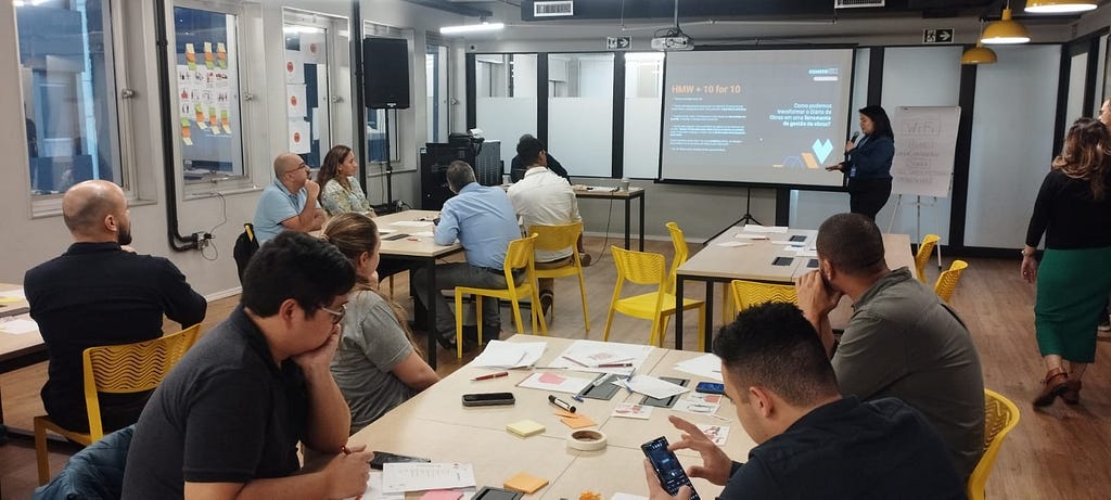 Um Designer facilitador apresentando em uma sala com microfone e datashow para diversos clientes, sentados na mesa, sobre o que iriam fazer em seguida.