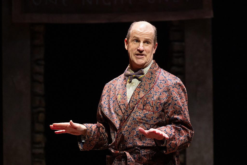 A man in a bowtie and patterned smoking jacket speaks with his hands out.