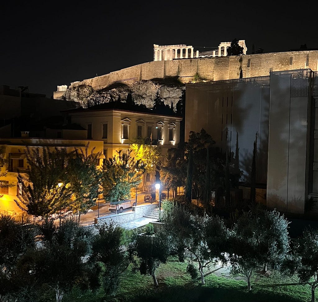 An image of the Acropolis in Athens, Greece at night, highlighting the Parthenon lit up with a dark sky and foreground.
