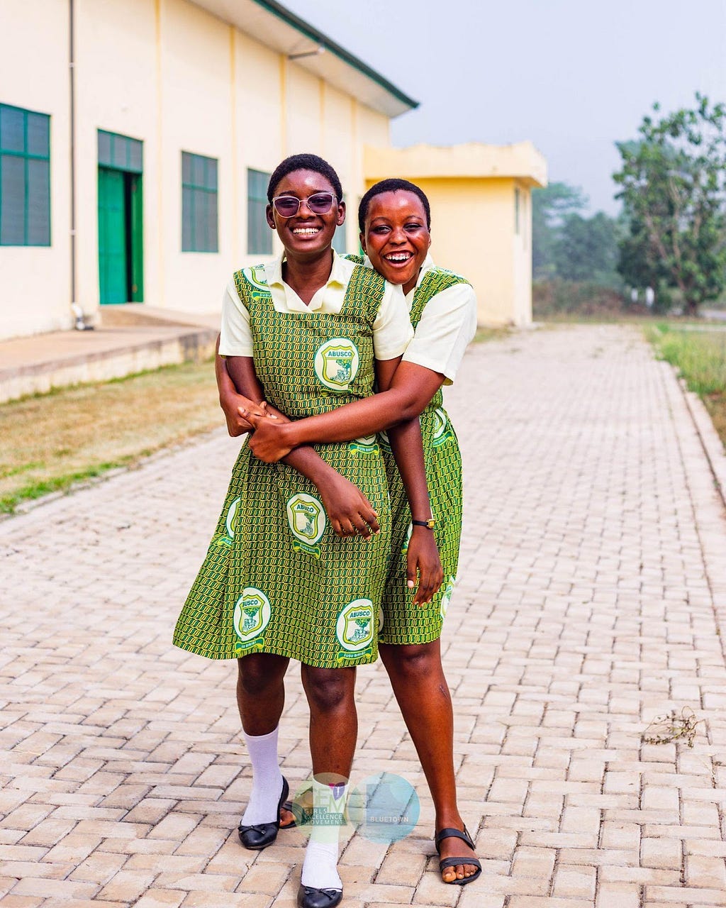 Two students dressed in matching green dresses smile and pose together in an embrace.