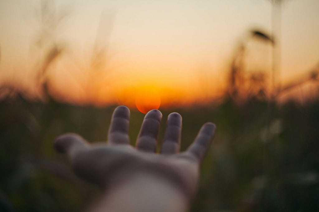 n out-of-focus background with a hand reaching out in the foreground representing help. There’s a beautiful sunset in the background.