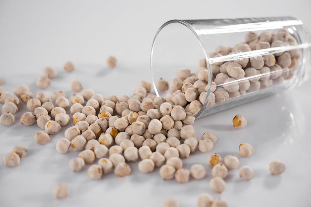 Dry, uncooked chickpeas in a glass jar, halfway poured out onto a white countertop.