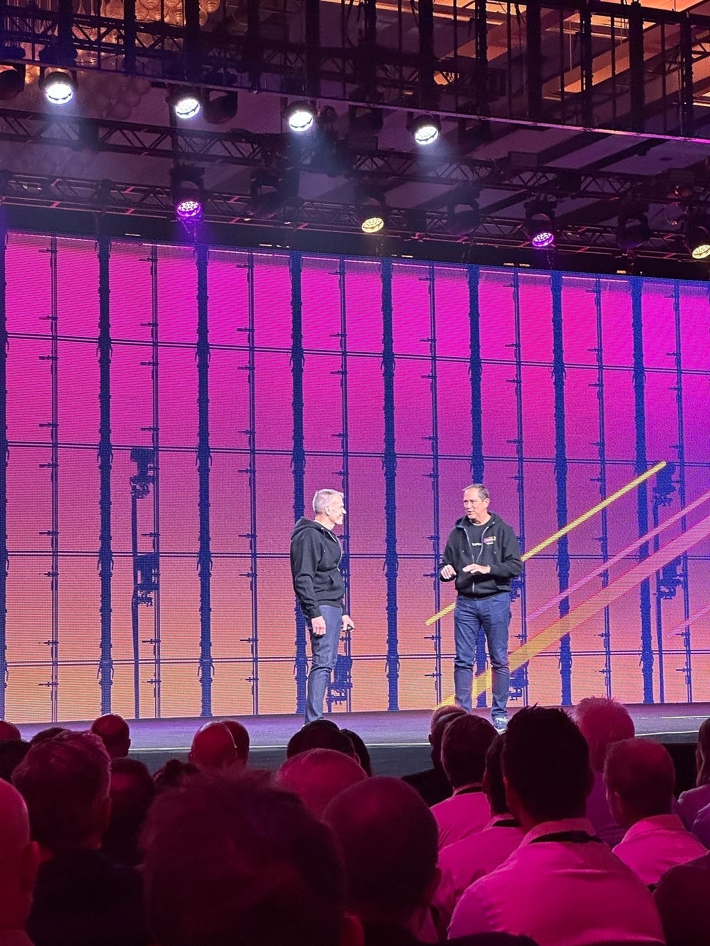 Two individuals are standing on a stage with a large screen behind them, speaking to an audience.