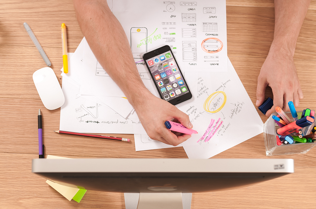 Person drawing a website wireframe on a desk with papers, pens, and a computer