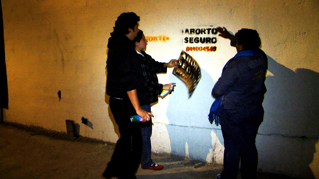 três pessoas pintam uma parede branca com a frase “aborto seguro” seguido de um número telefônico