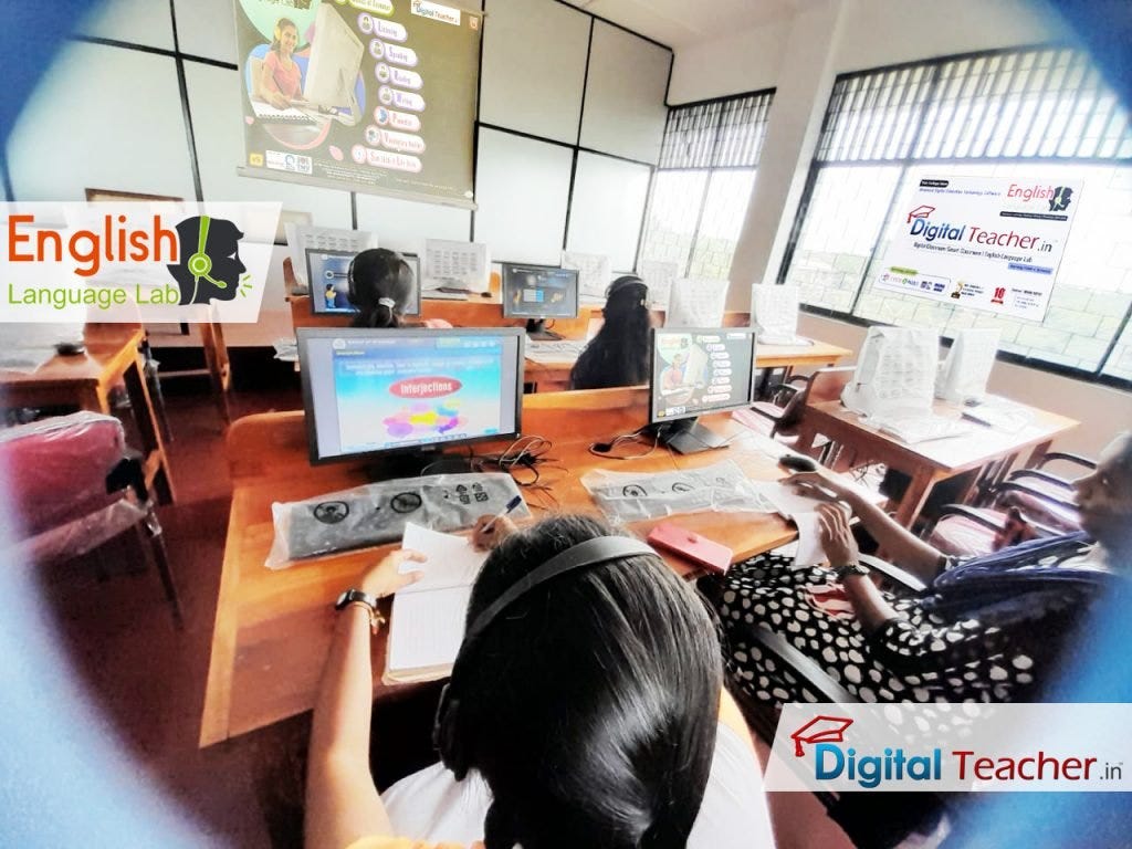 Students sit at computer equipment in an English language laboratory, with a large screen showing the ‘English Language Lab’ and a teacher nearby.