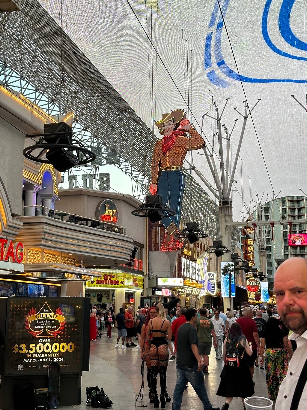 A busy street scene features a lit-up cowboy sign, various shops, a large digital advertisement, and a crowd of people, one person wearing an attention-grabbing outfit.