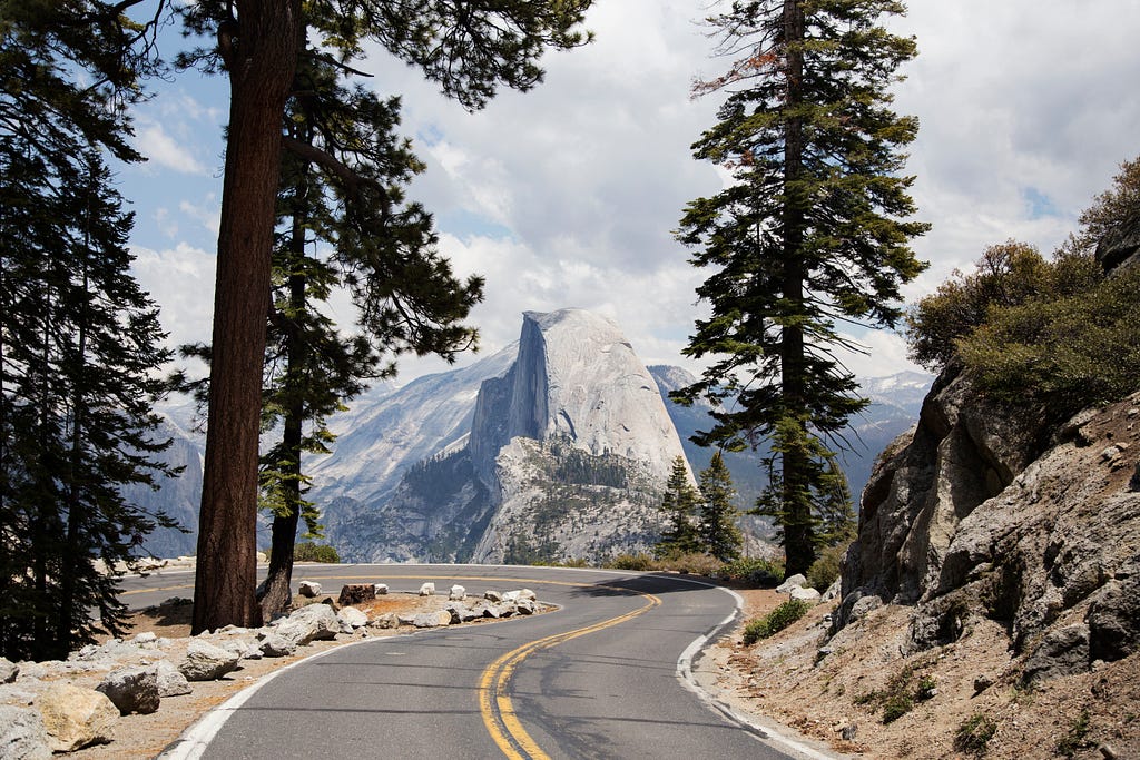 A photo of a road curving up with a view of the mountains overlooking trees and cliffs.