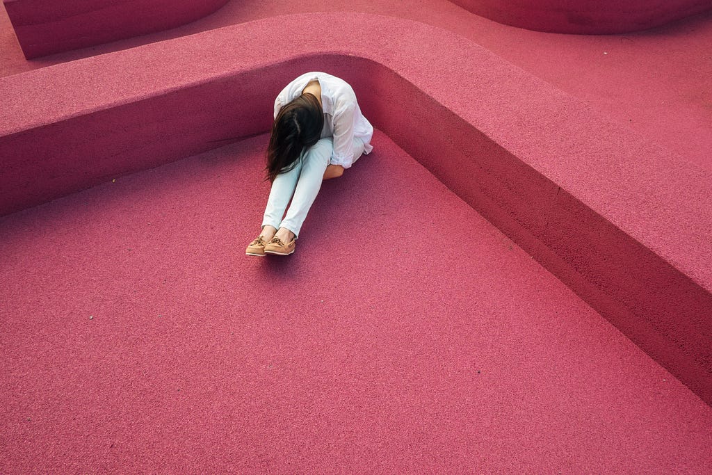 A person with long hair wearing a white shirt and light blue jeans sits with their head to their knees. Dark pink background.