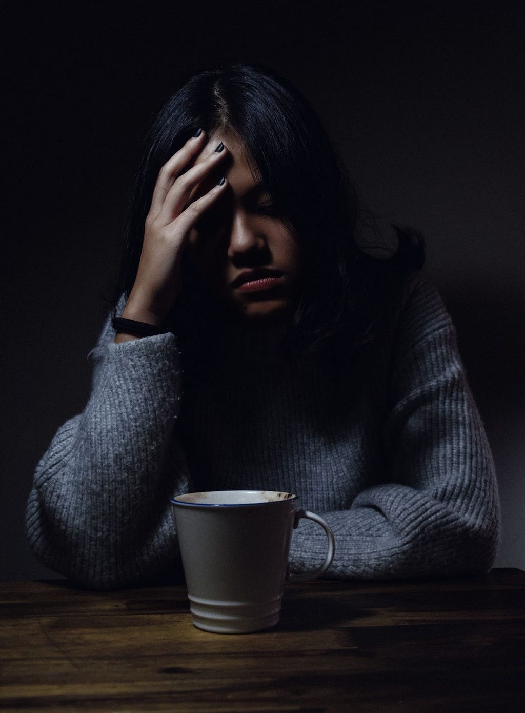 A woman sitting besides a table touching her forehead with closed eyes and a cups of coffee in front of her on the table.