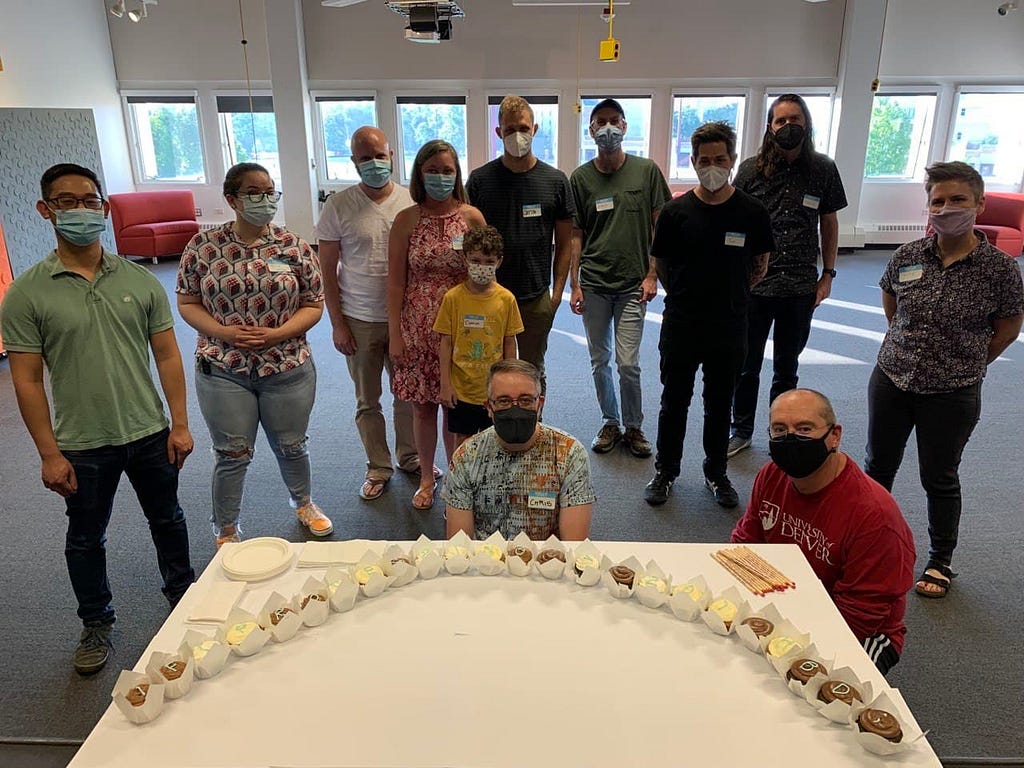 Photo of 12 attendees to the event standing and crouching behind a table displaying cupcakes arranged in the shape of an arc.