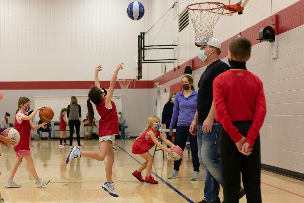 A photo of three girls dribbling and shooting basketballs as three coaches look on at the indoor basketball court.