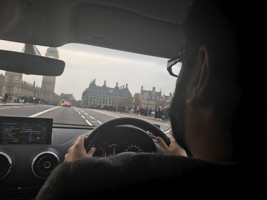 Over the shoulder view of bearded and bespectacled driver with hands on steering wheel. Big Ben visible ahead.