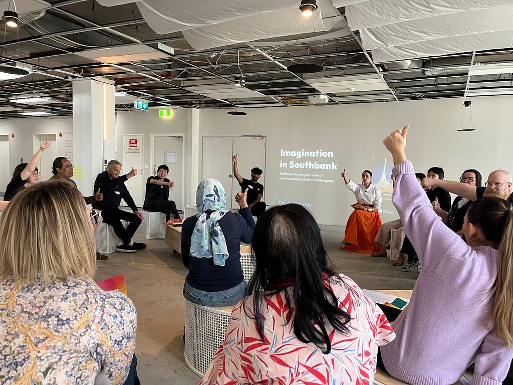 A group of people sit in a circle on stools with their thumbs up in the air, conveying that they think something is true.