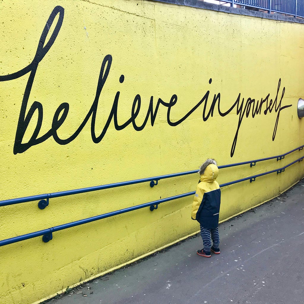 This image depicts a boy in a black and yellow jacket staring at a concrete wall that is painted yellow with the words “Believe in yourself.” written on it.