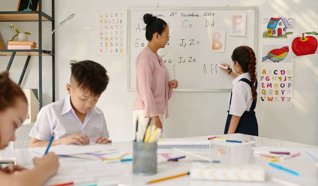 Teacher and child standing in front of a white board while two other children work at a table