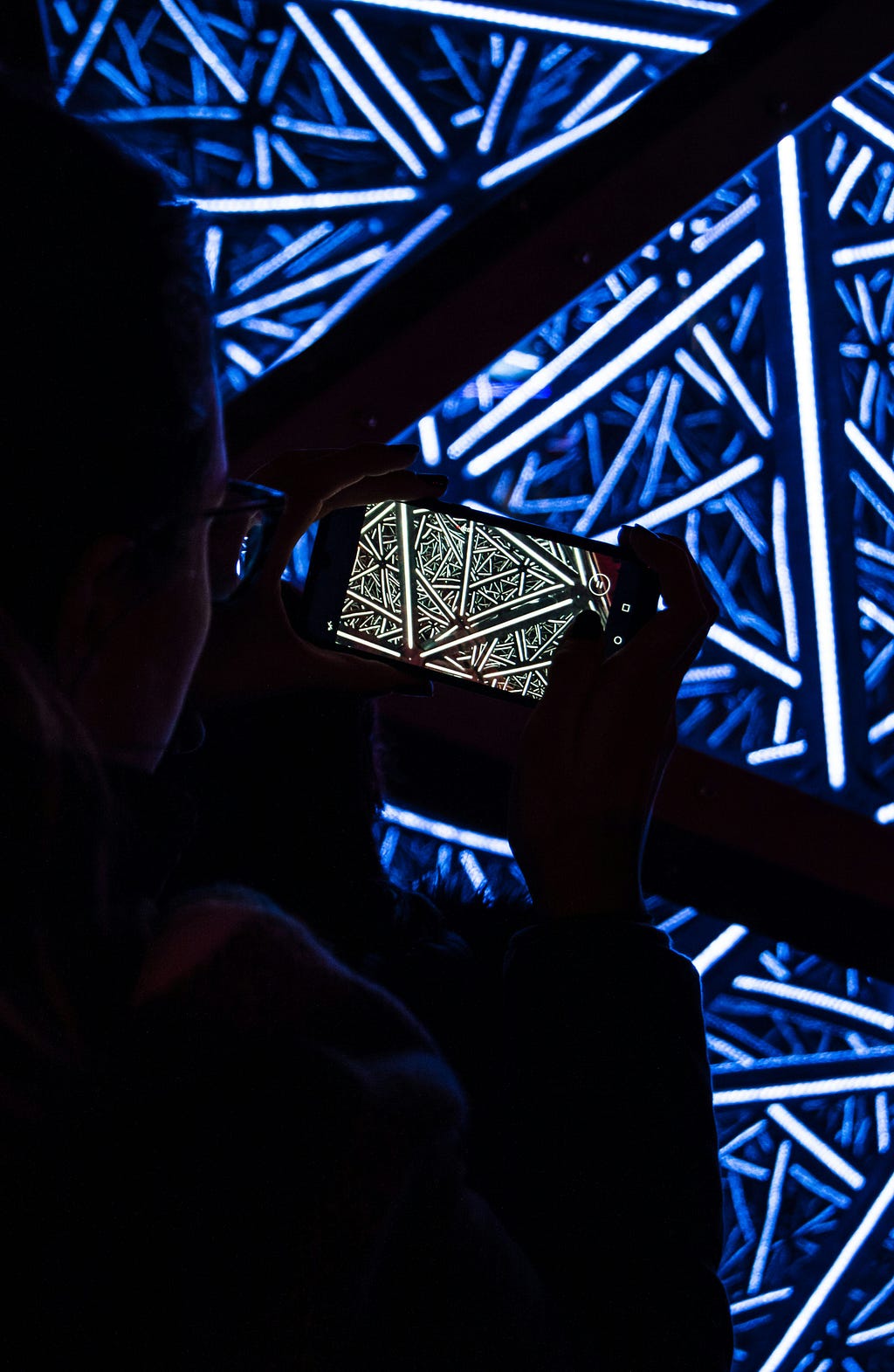 Person taking a photo of a bright blue light installation with intricate geometric patterns, reflected on the smartphone screen.