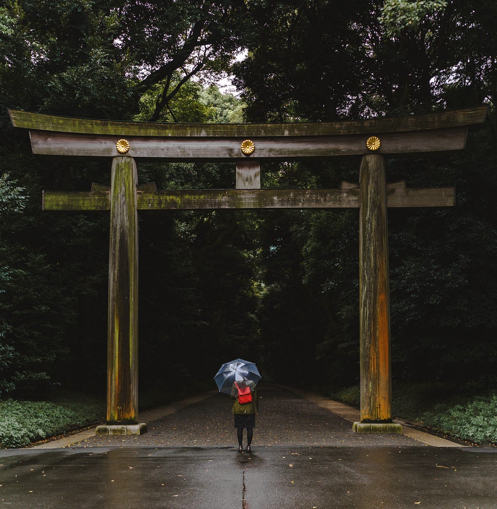 A woman with her back to the camera stands before a huge wooden torii gate with three golden chrysanthemum emblems stamped across its arch