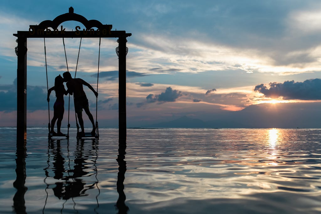 Two people kissing, during the sunset, standing on swings over the water.