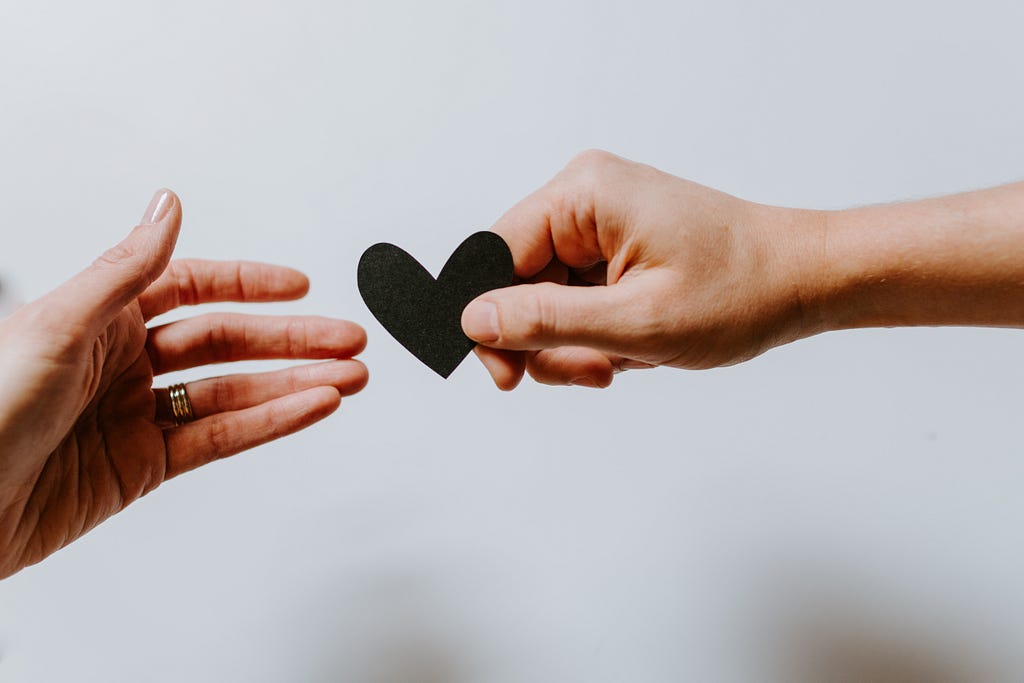 Two hands reaching towards each other. One is holding a black paper heart in the process of handing it off to the other hand.