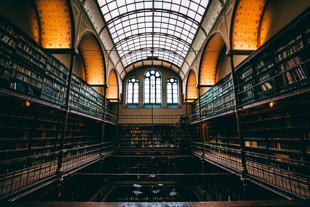 A library filled with bookcases of books on either side. A glass ceiling, criss-crossed with frames lets in a little light.