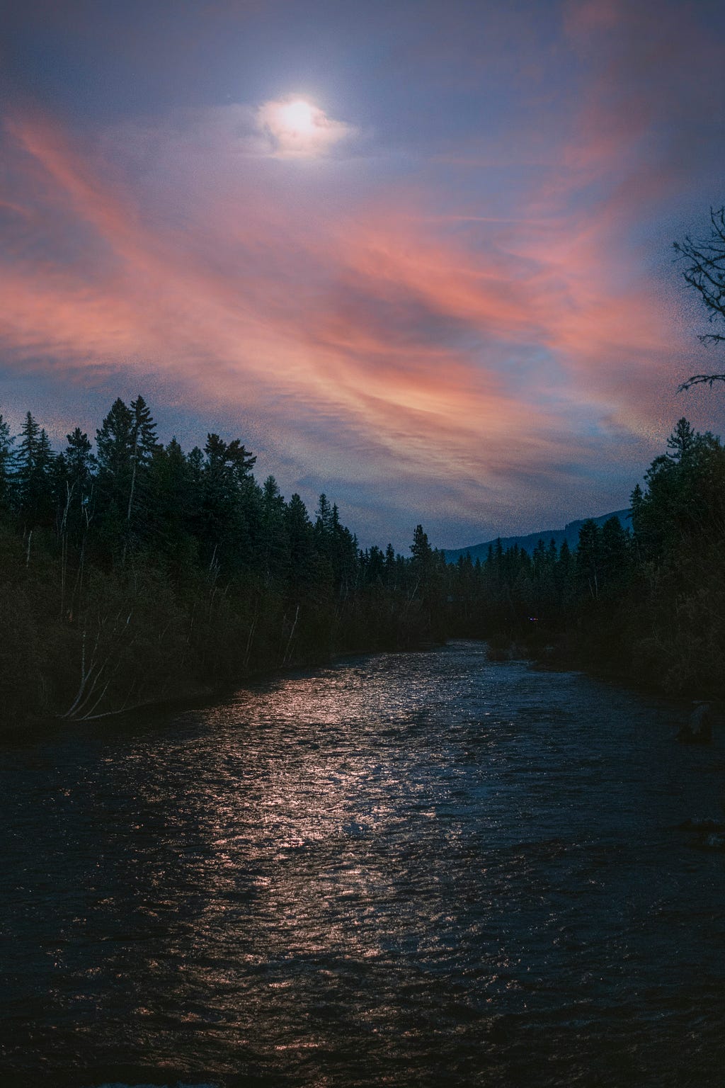 In the center is a river rushing, lots of ripples. Trees line each side of it tall trees with robust foliage. The moon is full and is shining through wispy sunset clouds and reflecting on the river. The clouds are pink with dark blue sky behind.