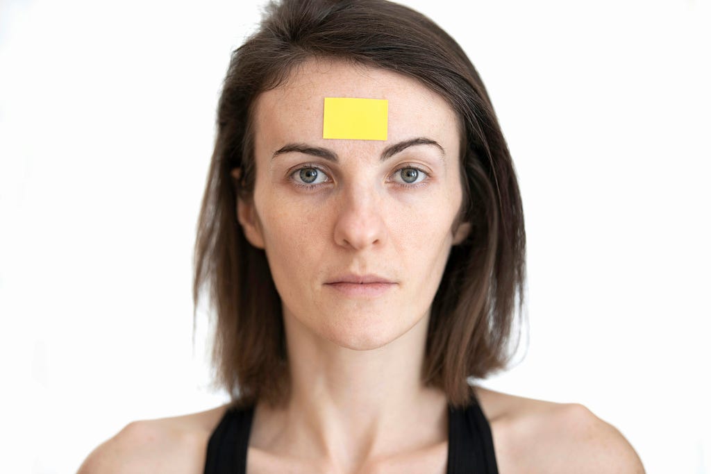 Head and shoulders of a woman looking at the camera. The woman has a yellow piece of paper stuck to her forehead.