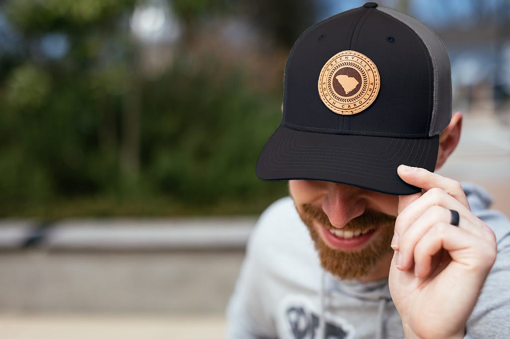 A bearded white man holds the brim of his baseball cap, which covers his eyes. The cap is black and gray with a tan, circular leather patch in the center that says “Greenville, South Carolina” and an image of the state of South Carolina.