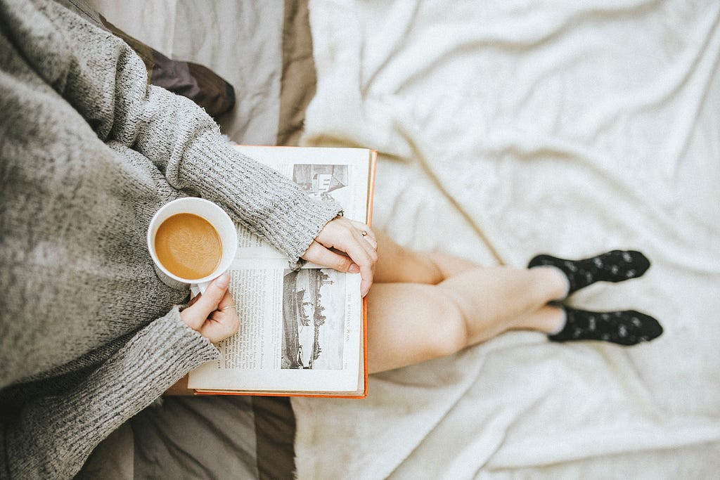 A birds-eye view of a person sitting with their feet crossed. They are holding a cup of coffee over an open book.