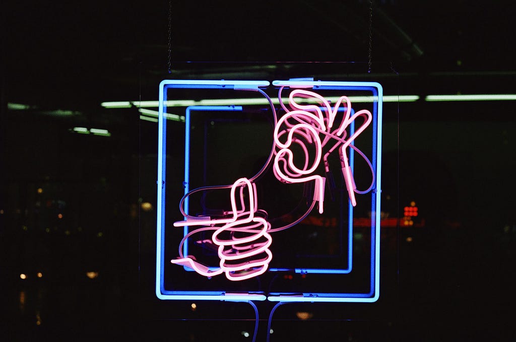 A blue square neon sign with 2 pink hands. One hand is holding the “thumbs up” gesture, the other holding the “okay” gesture.