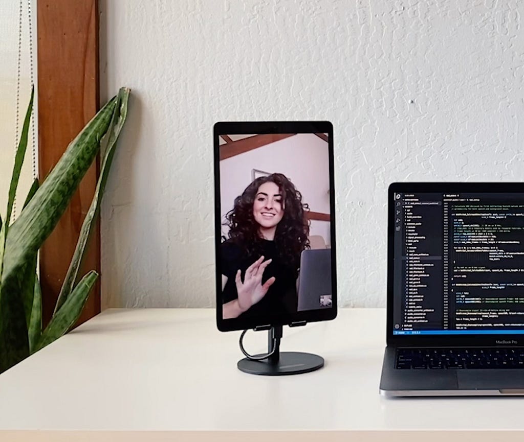 A photo of a laptop with a tablet on a stand next to it. The tablet shows a video-chat with a woman with her own laptop, waving at the camera.