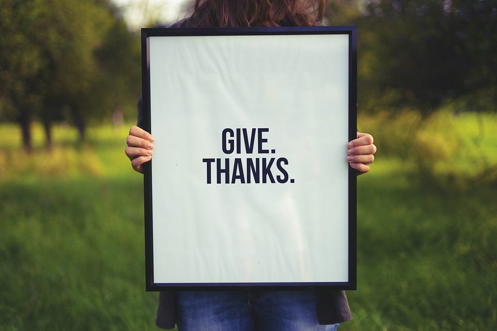 A person holding a sign with black margins and with white background that reads in black letters “GIVE.THANKS.”