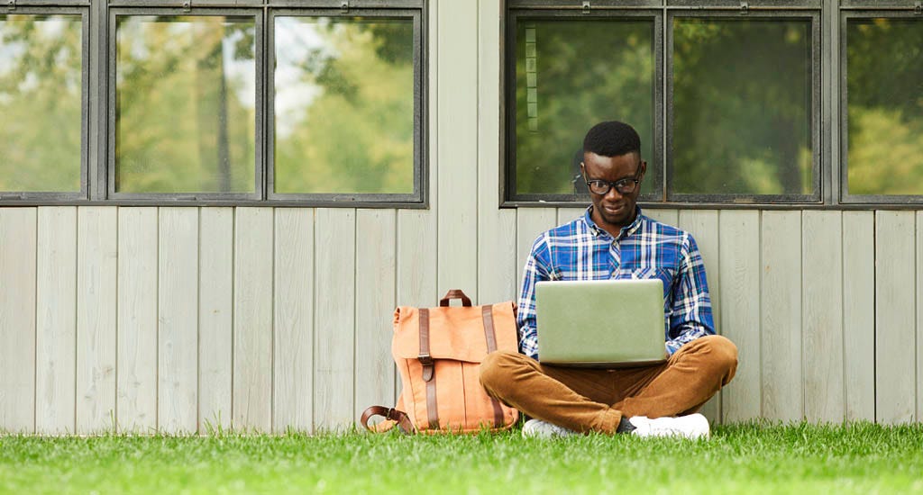 International college student entrepreneur using laptop outdoors while sitting in grass