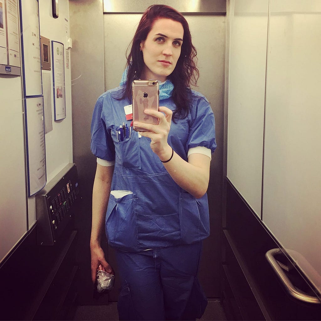Mirror selfie taken in hospital elevator, wearing scrubs.