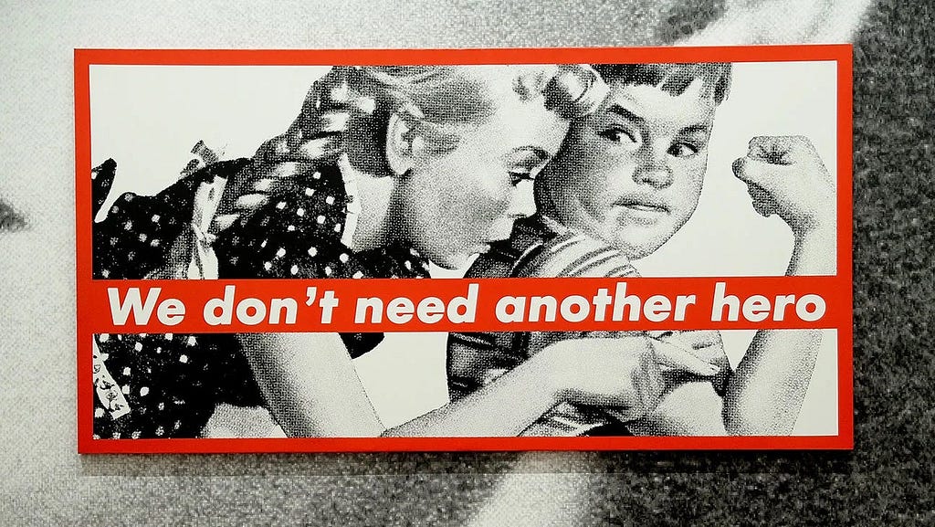 Barbara Kruger’s large black and white illustration of two kids in 5’s advertising style looking a their arm muscles — with red text banner splashed across reading: We don’t need another hero.