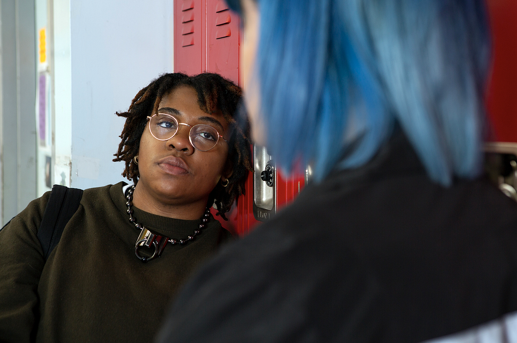 Picture of non-binary people talking next to locker rooms. One person is facing the camera, wearing a necklace and the other