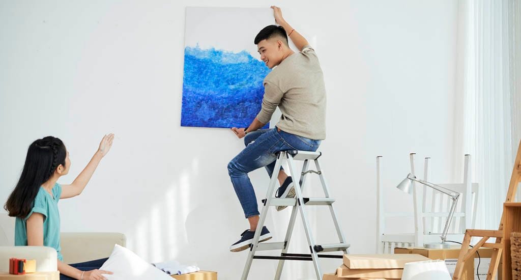 Woman guiding man on step ladder where to hang painting